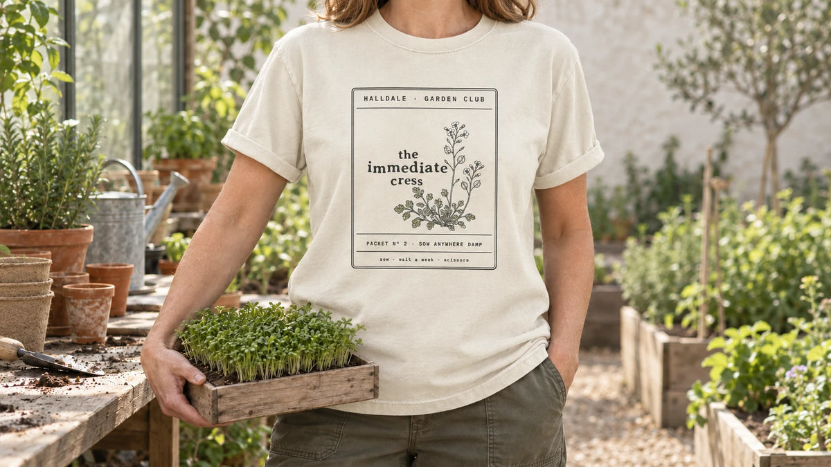 A woman in the Ivory Immediate Cress tee stands by a greenhouse potting bench with seed trays and terracotta pots, holding a shallow wooden seed tray of fresh green cress.
