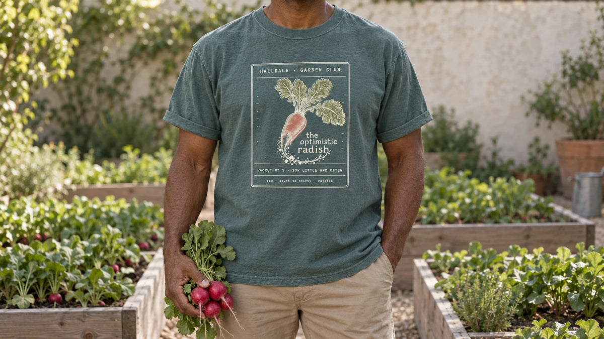 A man in the Blue Spruce Optimistic Radish tee stands by raised vegetable beds with neat rows of radish leaves, holding a bunch of fresh red radishes with their leafy tops.