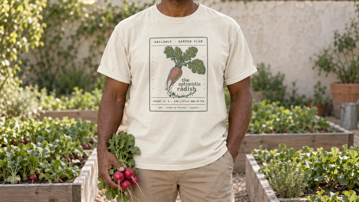 A man in the Ivory Optimistic Radish tee stands by raised vegetable beds with neat rows of radish leaves, holding a bunch of fresh red radishes with their leafy tops.
