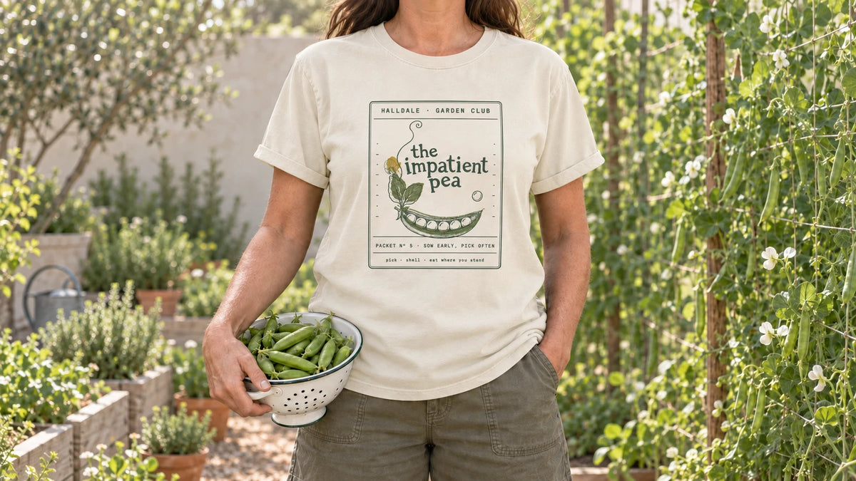 A woman in the Ivory Impatient Pea tee stands by a vegetable garden beside a pea trellis, holding a small enamel colander of fresh pea pods.