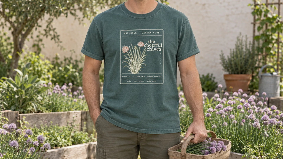 A man in the Blue Spruce Cheerful Chives tee stands by an herb bed full of flowering chives with lilac pompom flowers, holding a small wooden trug of cut chives with a few lilac flowers.