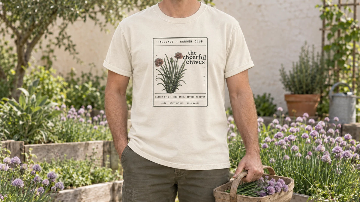 A man in the Ivory Cheerful Chives tee stands by an herb bed full of flowering chives with lilac pompom flowers, holding a small wooden trug of cut chives with a few lilac flowers.
