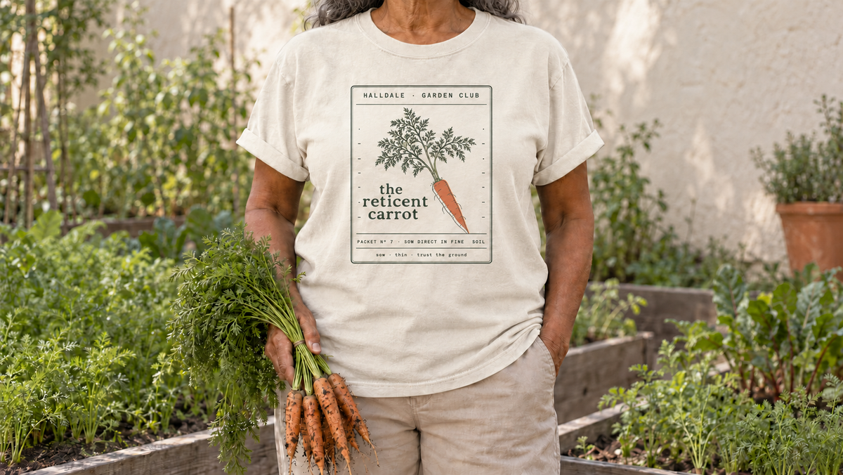 A woman in the Ivory Reticent Carrot tee stands by raised beds of fine dark soil with feathery carrot tops, holding a bunch of freshly pulled carrots with feathery green tops.