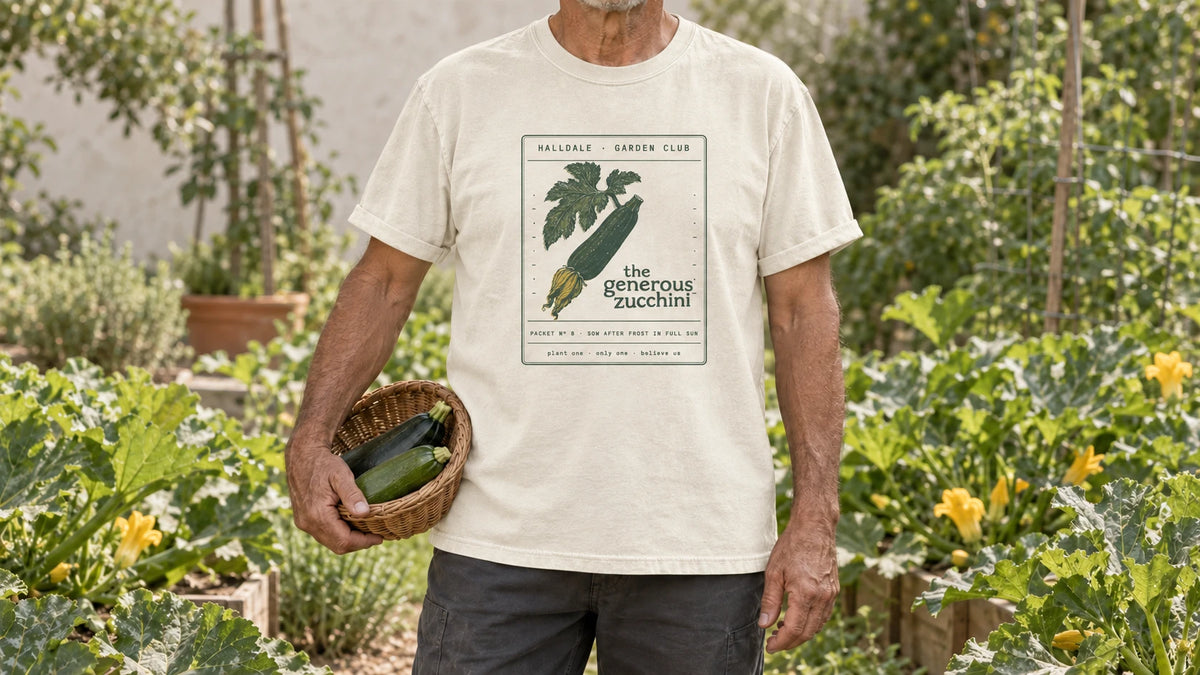 A man in the Ivory Generous Zucchini tee stands by a vegetable bed of sprawling zucchini plants with broad leaves and yellow flowers, holding a shallow wicker basket holding two zucchini.