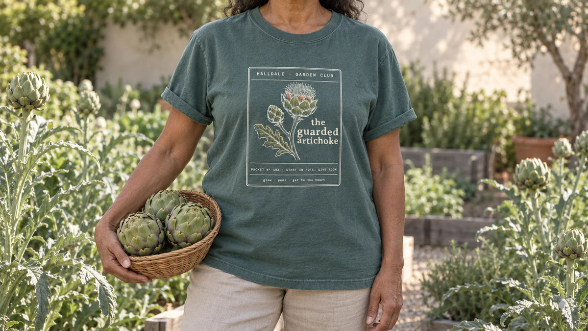 A woman in the Blue Spruce Guarded Artichoke tee stands by tall silvery architectural artichoke plants, holding a shallow wicker basket of three green artichokes.