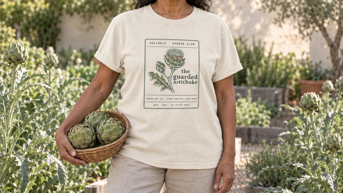 A woman in the Ivory Guarded Artichoke tee stands by tall silvery architectural artichoke plants, holding a shallow wicker basket of three green artichokes.