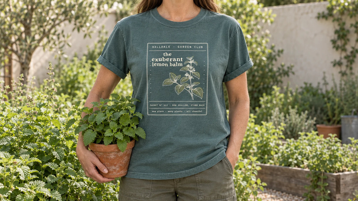 A woman in the Blue Spruce Exuberant Lemon Balm tee stands by an herb bed of lush lemon balm, holding a terracotta pot of lemon balm.