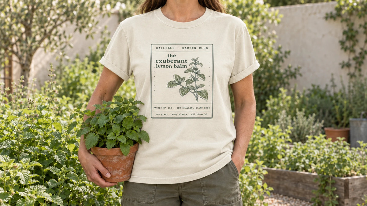 A woman in the Ivory Exuberant Lemon Balm tee stands by an herb bed of lush lemon balm, holding a terracotta pot of lemon balm.