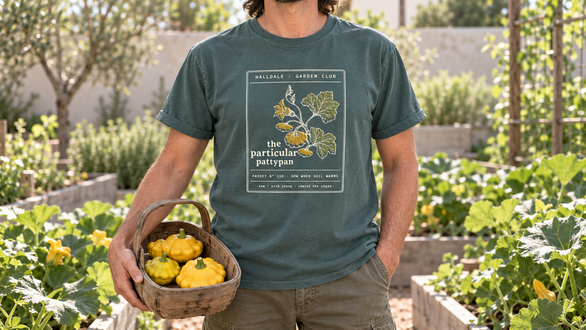 A man in the Blue Spruce Particular Pattypan tee stands in a summer squash garden, holding a wooden trug of yellow pattypan squash.
