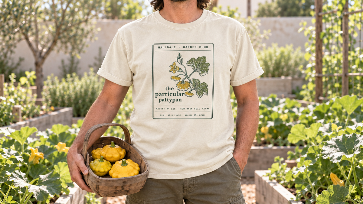 A man in the Ivory Particular Pattypan tee stands in a summer squash garden, holding a wooden trug of yellow pattypan squash.