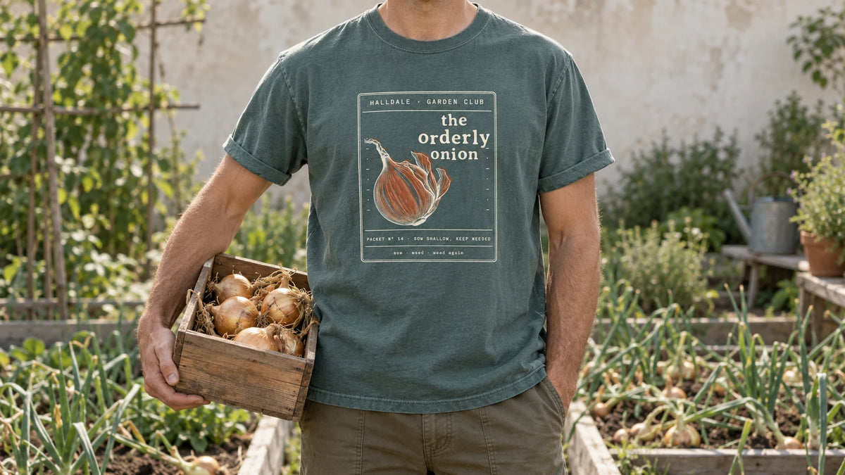A man in the Blue Spruce Orderly Onion tee stands by an onion bed with tidy rows and a few flopped green tops, holding a shallow wooden crate of cured golden onions.