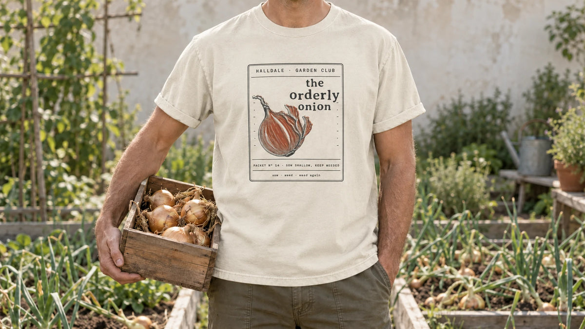 A man in the Ivory Orderly Onion tee stands by an onion bed with tidy rows and a few flopped green tops, holding a shallow wooden crate of cured golden onions.