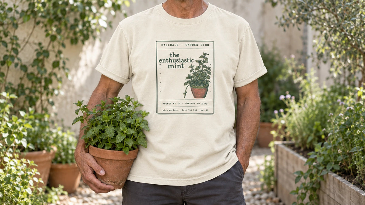 A man in the Ivory Enthusiastic Mint tee stands by a shady garden corner with mint spilling out of terracotta pots, holding a terracotta pot of bright green mint.