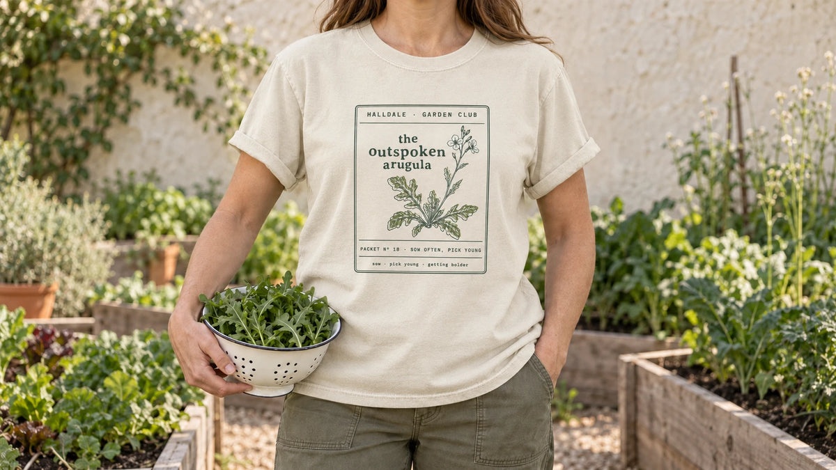 A woman in the Ivory Outspoken Arugula tee stands by raised salad beds of young leafy greens, holding a small enamel colander of fresh arugula leaves.