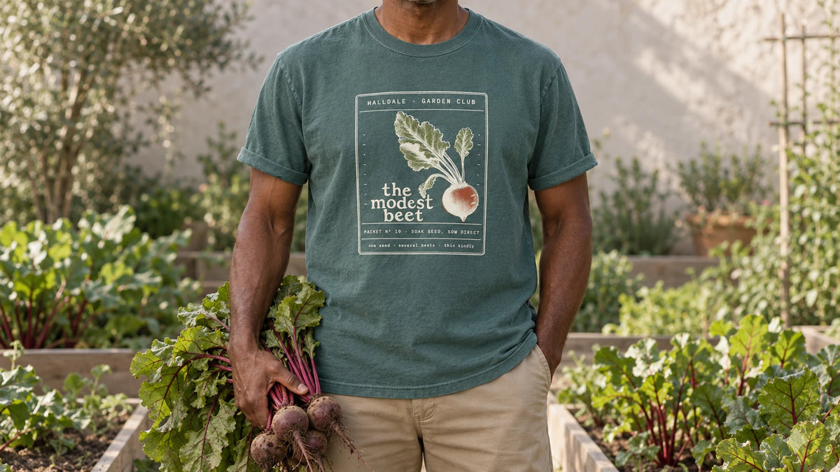 A man in the Blue Spruce Modest Beet tee stands by raised beds of beets with red-veined leaves, holding a bunch of freshly pulled beets with red-veined leaves.