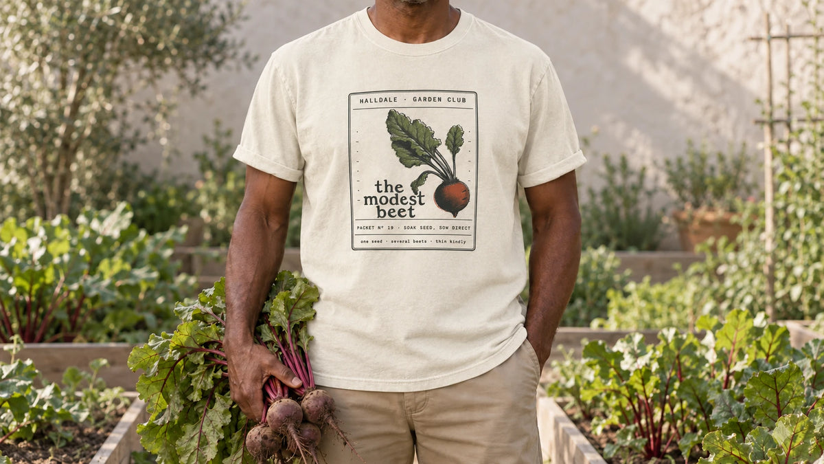 A man in the Ivory Modest Beet tee stands by raised beds of beets with red-veined leaves, holding a bunch of freshly pulled beets with red-veined leaves.