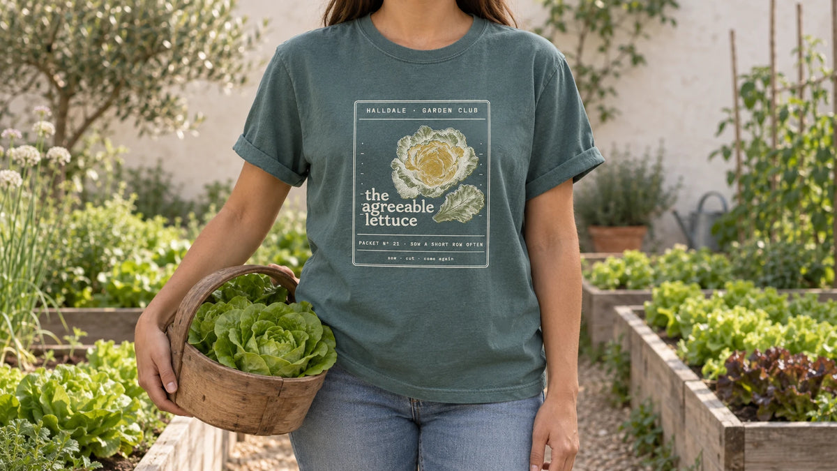 A woman in the Blue Spruce Agreeable Lettuce tee stands by raised beds with neat rows of butterhead lettuce, holding a wooden trug holding two heads of butterhead lettuce.
