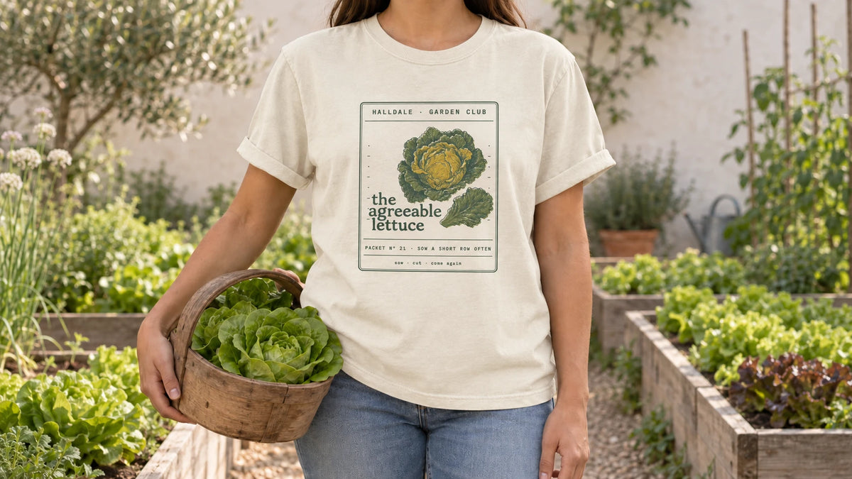 A woman in the Ivory Agreeable Lettuce tee stands by raised beds with neat rows of butterhead lettuce, holding a wooden trug holding two heads of butterhead lettuce.