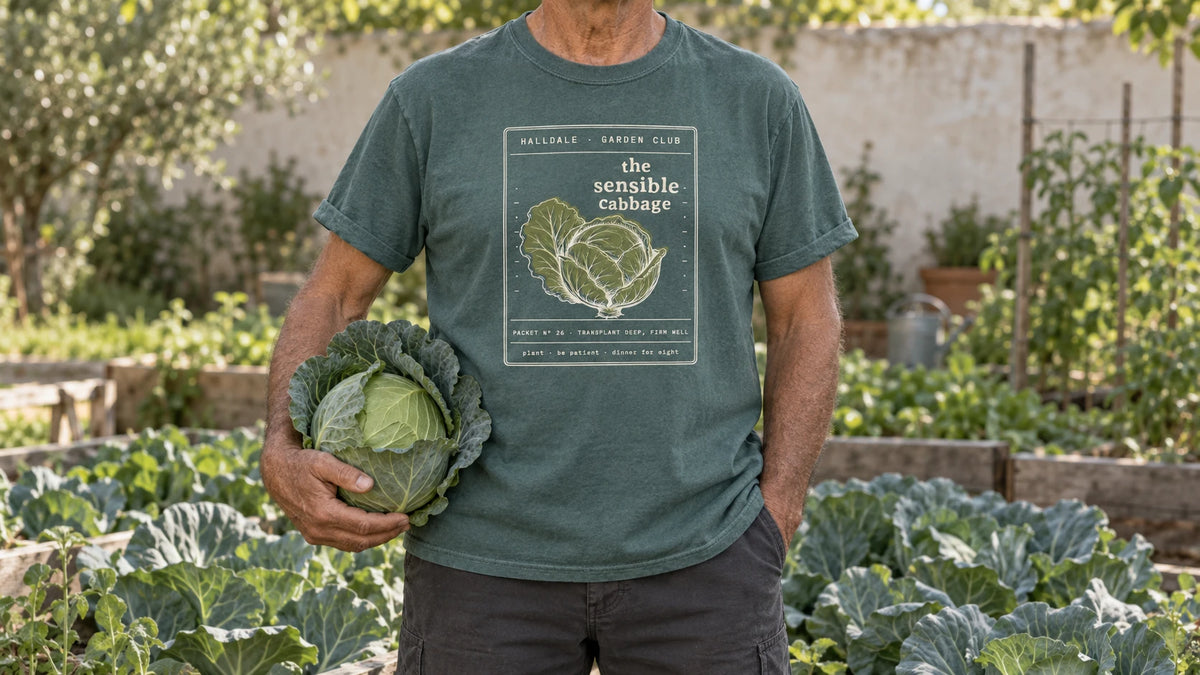 A man in the Blue Spruce Sensible Cabbage tee stands by a cabbage patch of large blue-green heads, holding one large green cabbage cradled against the hip.
