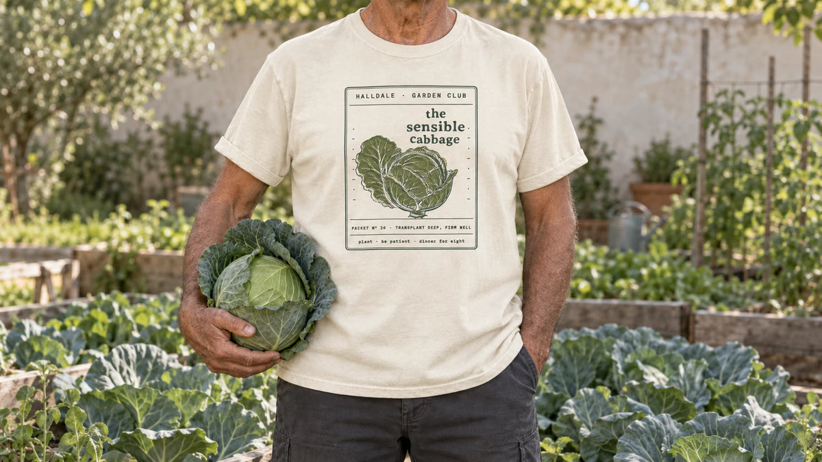 A man in the Ivory Sensible Cabbage tee stands by a cabbage patch of large blue-green heads, holding one large green cabbage cradled against the hip.