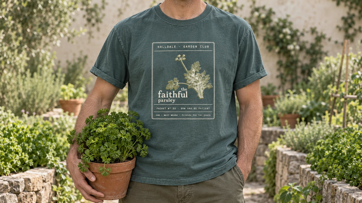 A man in the Blue Spruce Faithful Parsley tee stands by a stone-edged herb bed of curly parsley, holding a terracotta pot of curly parsley.