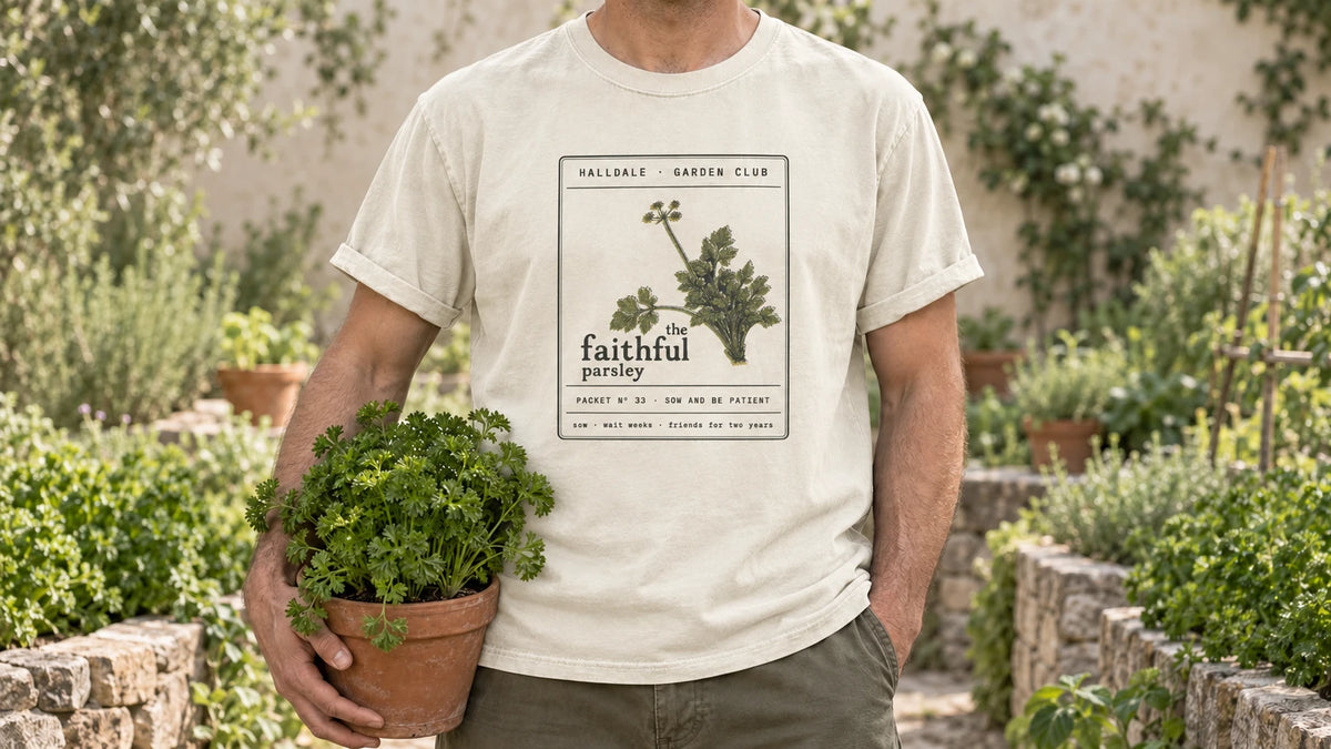 A man in the Ivory Faithful Parsley tee stands by a stone-edged herb bed of curly parsley, holding a terracotta pot of curly parsley.