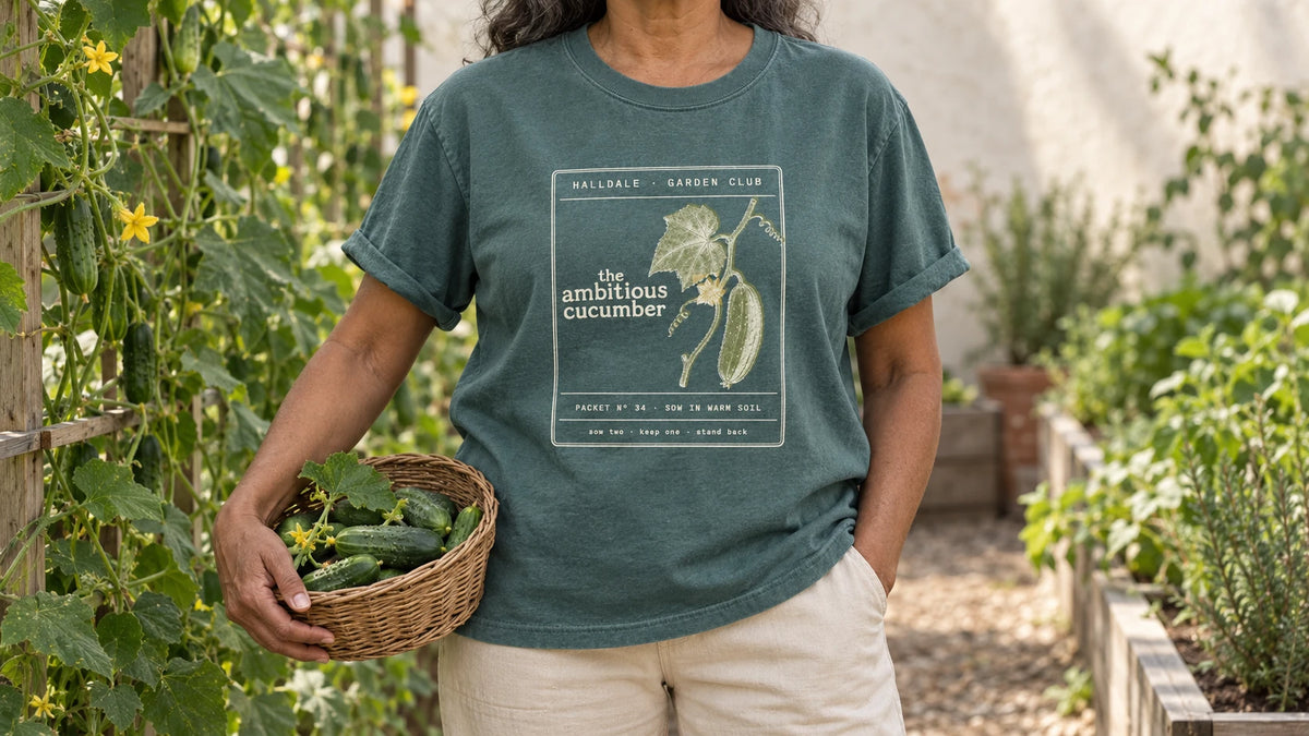 A woman in the Blue Spruce Ambitious Cucumber tee stands by cucumber vines climbing a wooden trellis, holding a shallow wicker basket of fresh cucumbers.