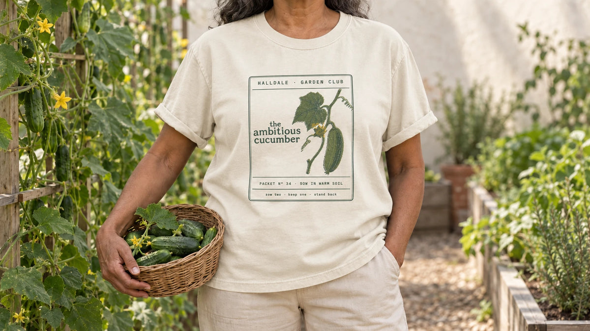 A woman in the Ivory Ambitious Cucumber tee stands by cucumber vines climbing a wooden trellis, holding a shallow wicker basket of fresh cucumbers.