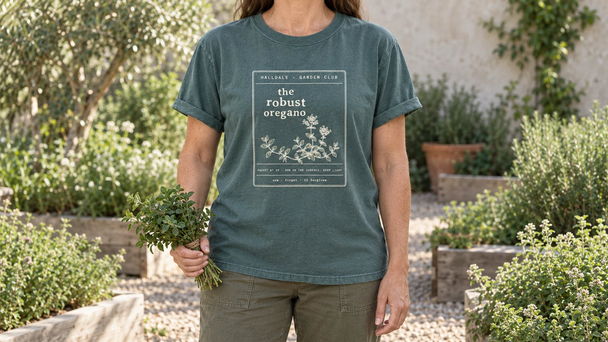 A woman in the Blue Spruce Robust Oregano tee stands by a sunny gravel herb garden with low mounds of oregano, holding a small bundle of fresh oregano sprigs tied with twine.