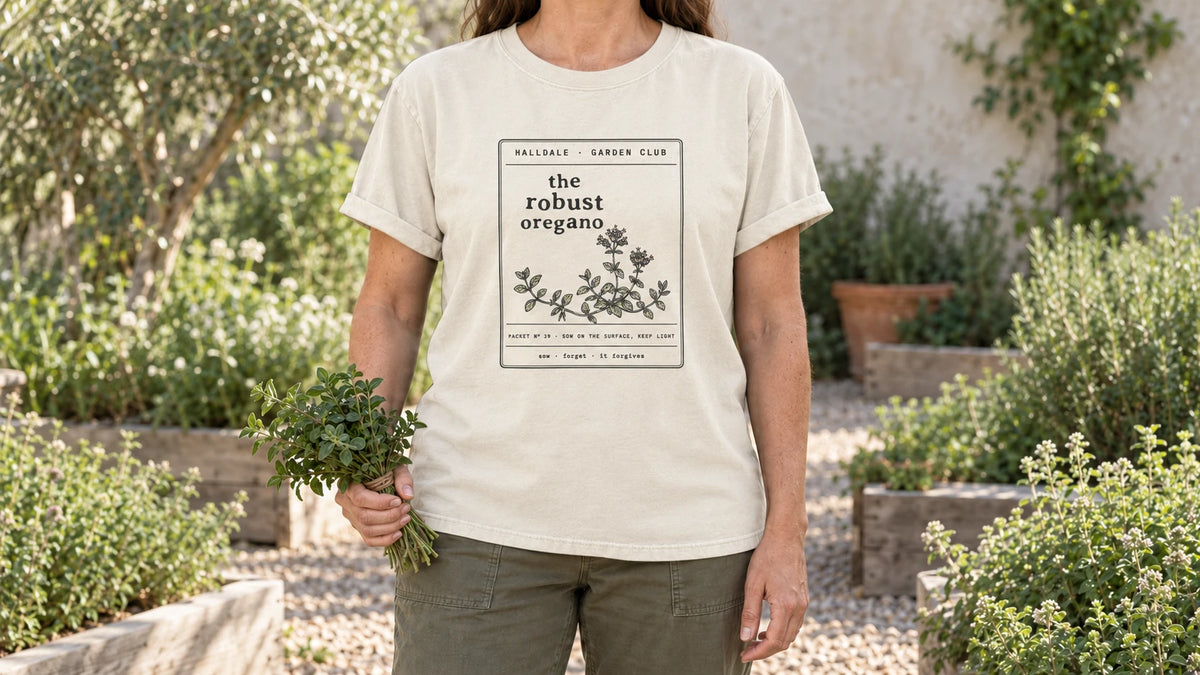 A woman in the Ivory Robust Oregano tee stands by a sunny gravel herb garden with low mounds of oregano, holding a small bundle of fresh oregano sprigs tied with twine.