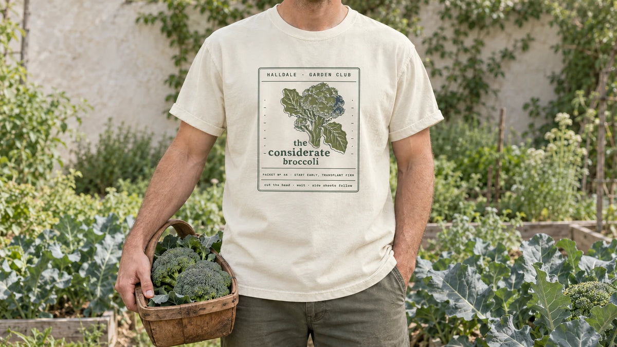 A man in the Ivory Considerate Broccoli tee stands by a brassica bed of broccoli plants with broad blue-green leaves, holding a wooden trug holding two fresh broccoli heads.