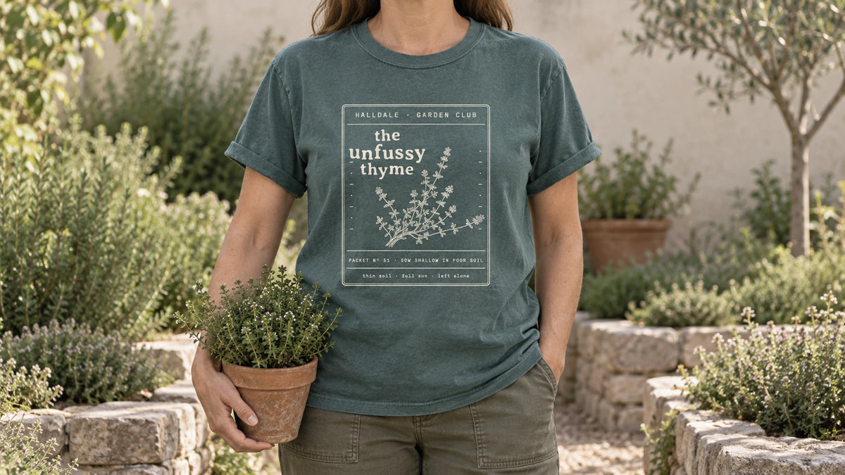 A woman in the Blue Spruce Unfussy Thyme tee stands by a stone-edged herb bed with low mounds of thyme, holding a small terracotta pot of thyme.