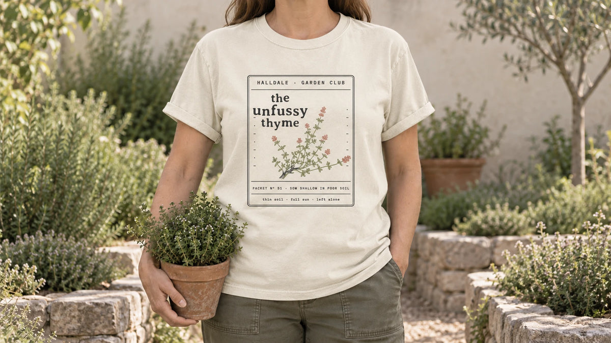A woman in the Ivory Unfussy Thyme tee stands by a stone-edged herb bed with low mounds of thyme, holding a small terracotta pot of thyme.