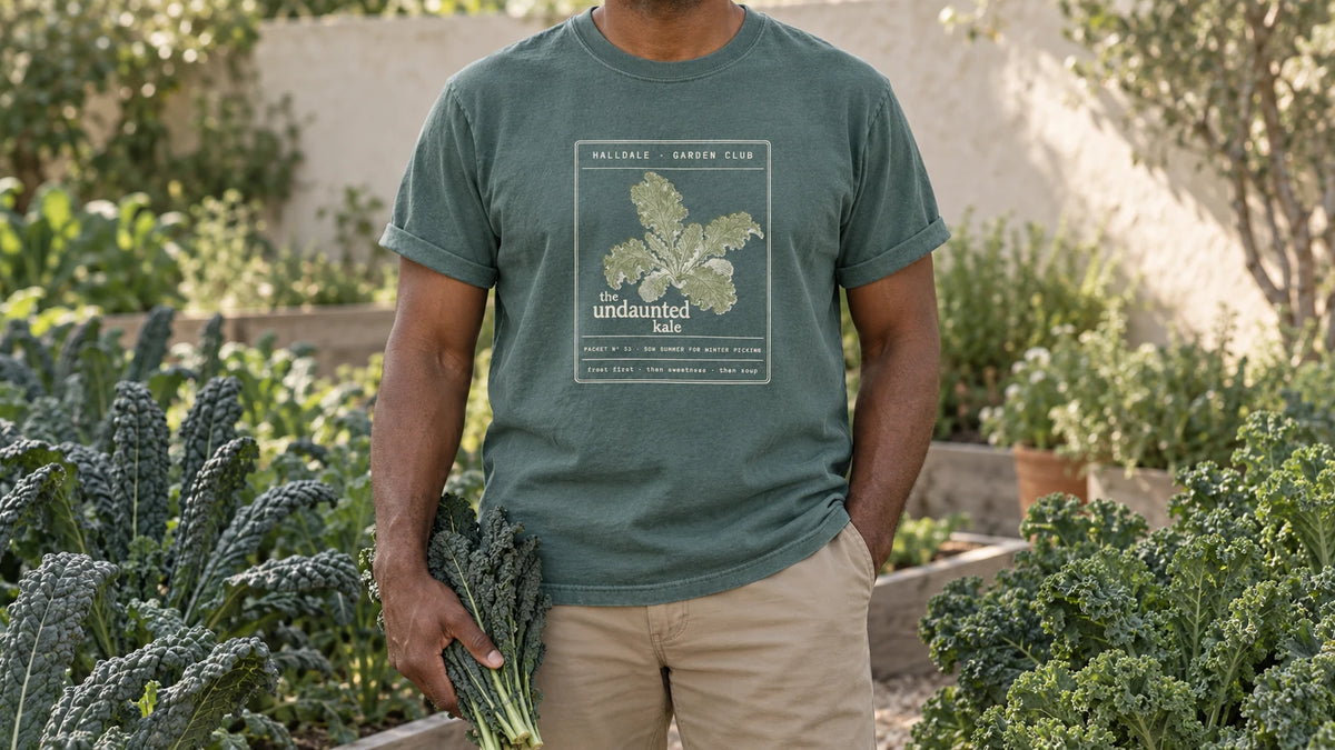 A man in the Blue Spruce Undaunted Kale tee stands by a kale bed of tall lacinato and curly kale, holding a bunch of dark lacinato kale leaves.