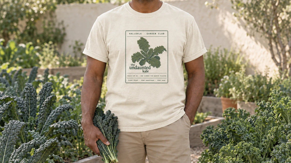 A man in the Ivory Undaunted Kale tee stands by a kale bed of tall lacinato and curly kale, holding a bunch of dark lacinato kale leaves.