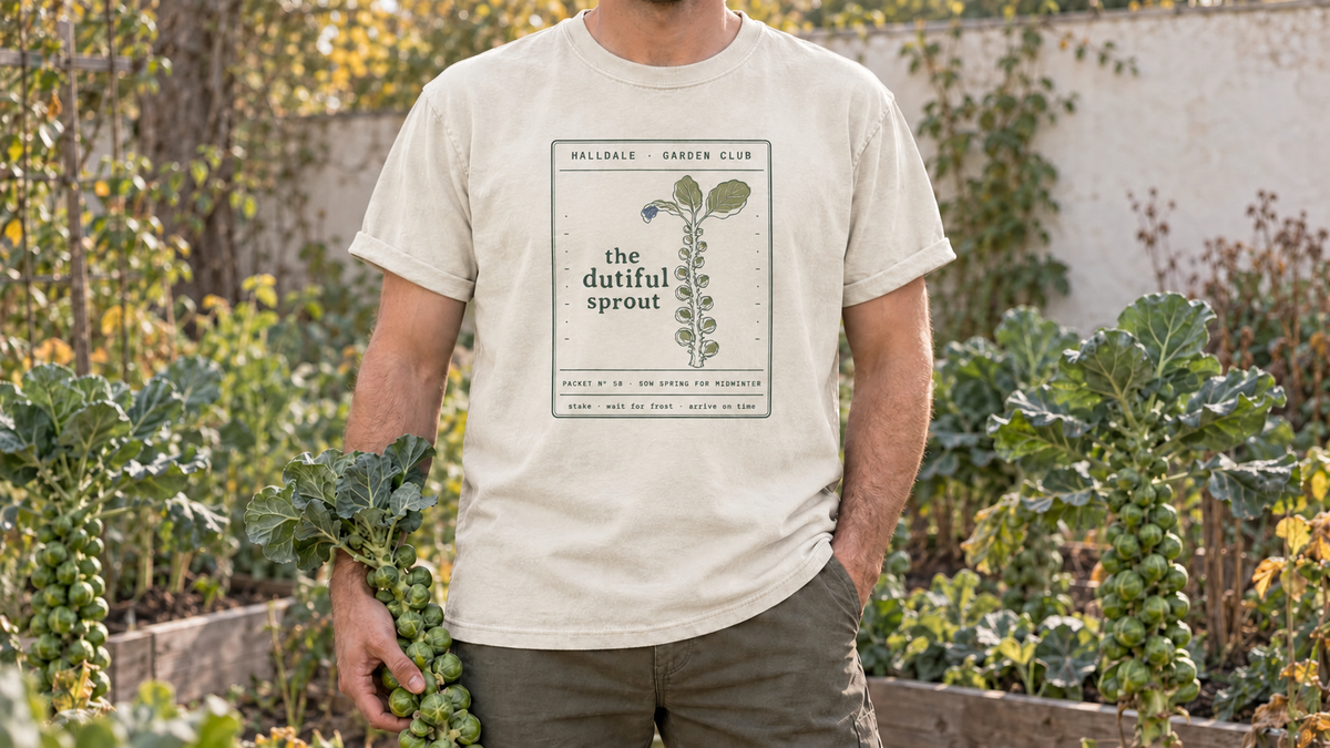 A man wearing the updated Dutiful Sprout tee in Ivory, holding a Brussels sprout stalk in a late fall brassica garden.