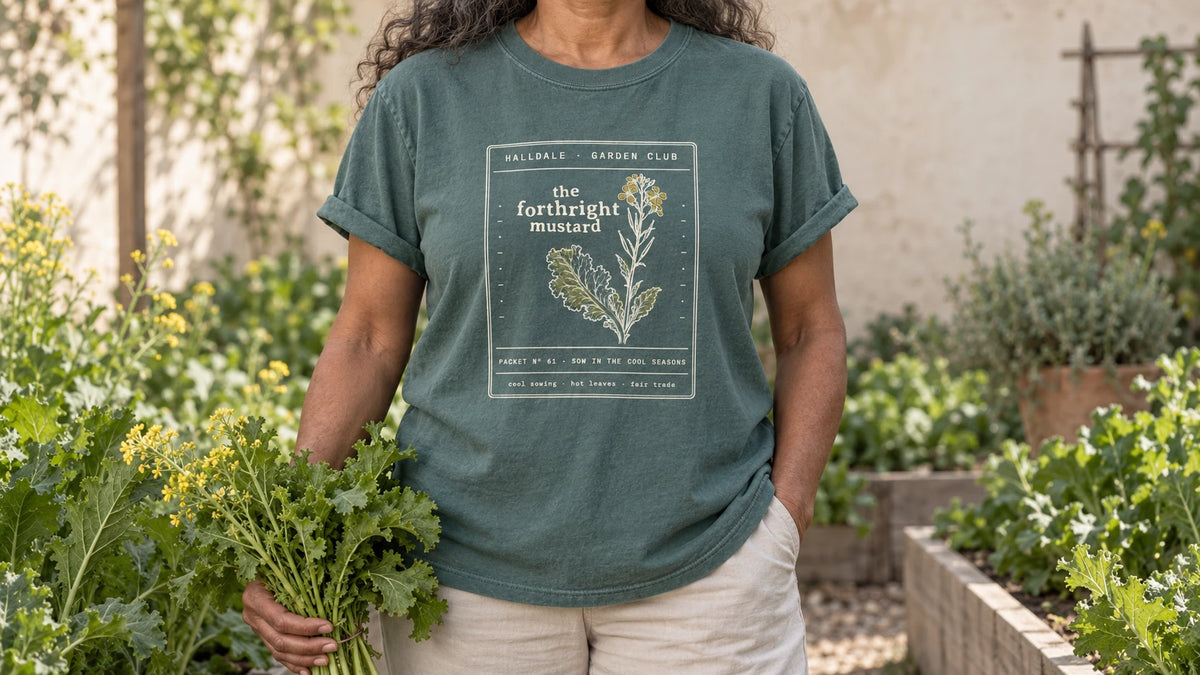 A woman in the Blue Spruce Forthright Mustard tee stands by a cool-season bed of mustard greens with a few yellow flowers, holding a bunch of frilly mustard greens.