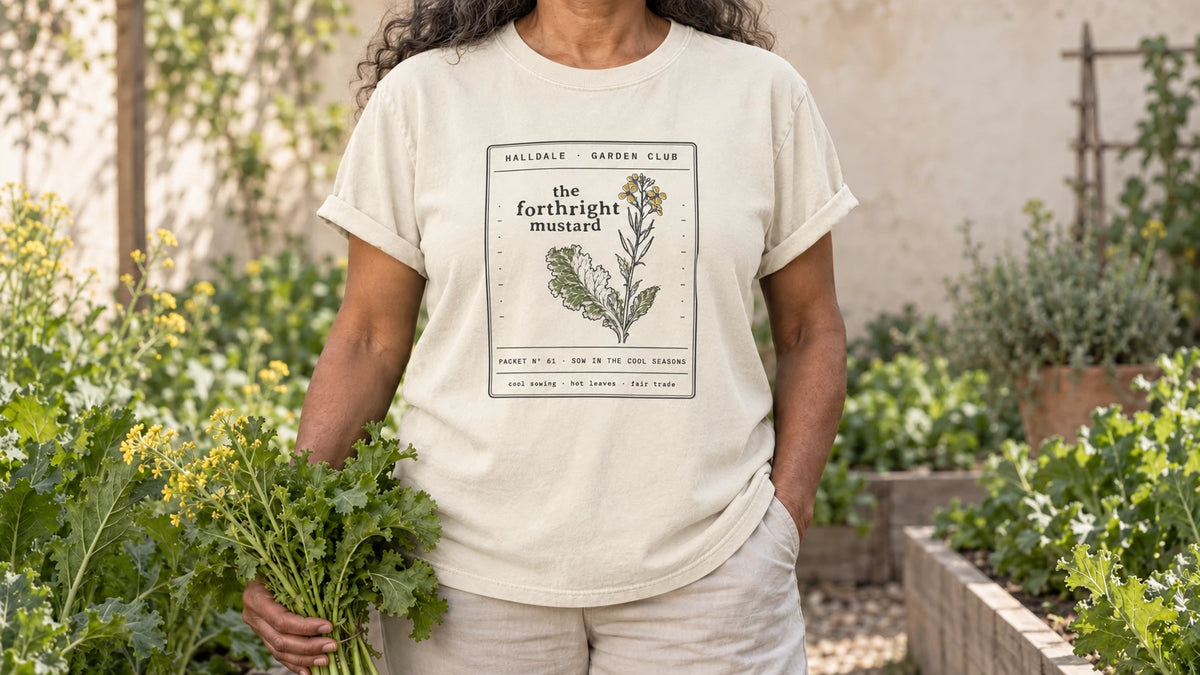 A woman in the Ivory Forthright Mustard tee stands by a cool-season bed of mustard greens with a few yellow flowers, holding a bunch of frilly mustard greens.