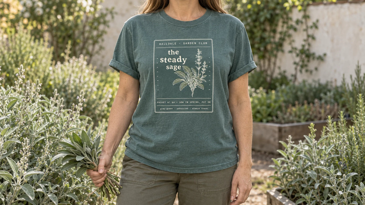 A woman in the Blue Spruce Steady Sage tee stands by an herb garden of silvery gray-green sage bushes, holding a small bundle of fresh sage leaves tied with twine.