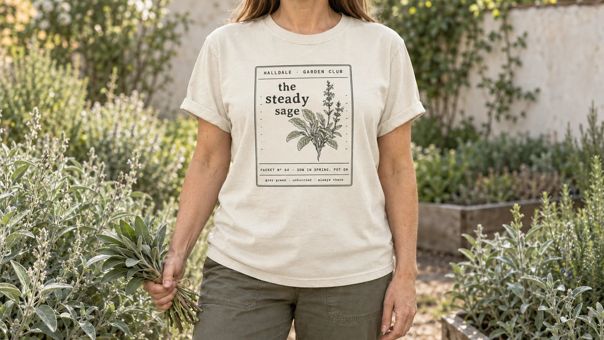 A woman in the Ivory Steady Sage tee stands by an herb garden of silvery gray-green sage bushes, holding a small bundle of fresh sage leaves tied with twine.