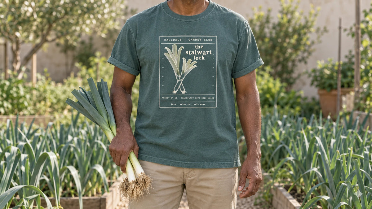 A man in the Blue Spruce Stalwart Leek tee stands by a bed of tall blue-green leeks, holding three fresh leeks.