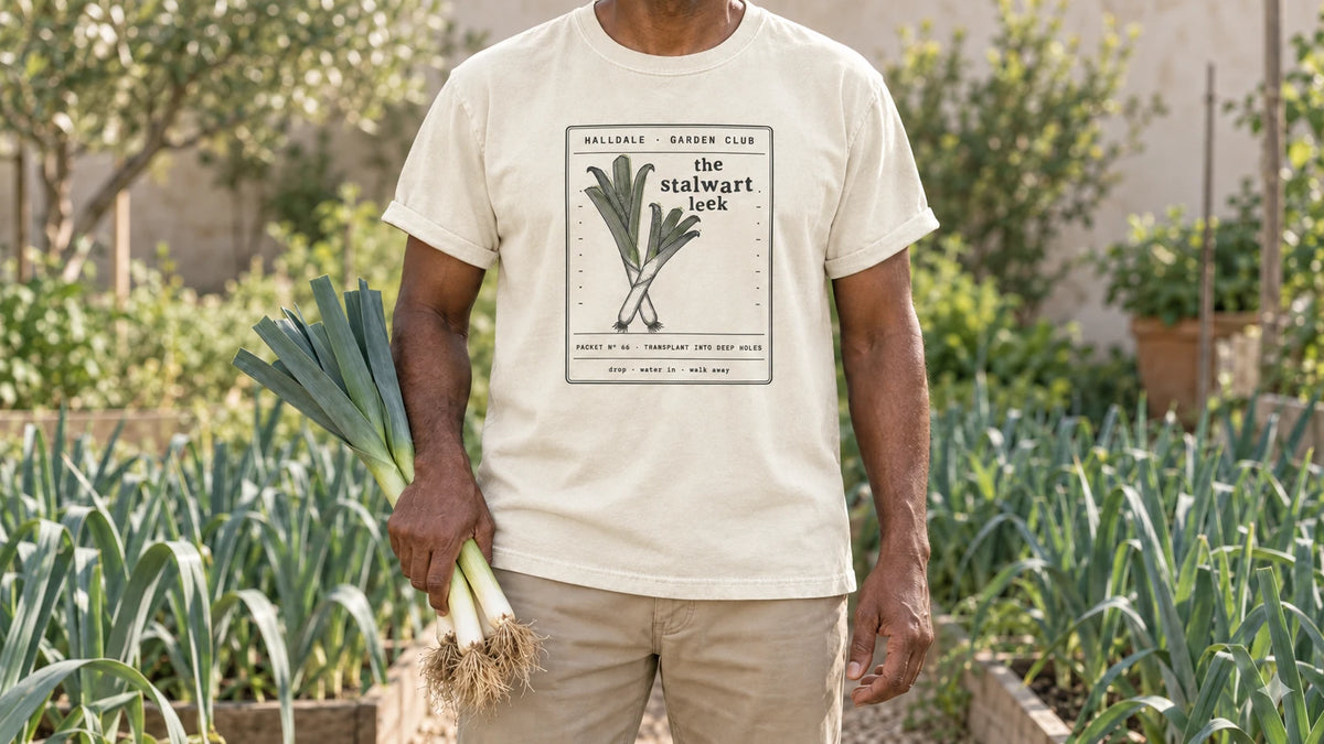 A man in the Ivory Stalwart Leek tee stands by a bed of tall blue-green leeks, holding three fresh leeks.
