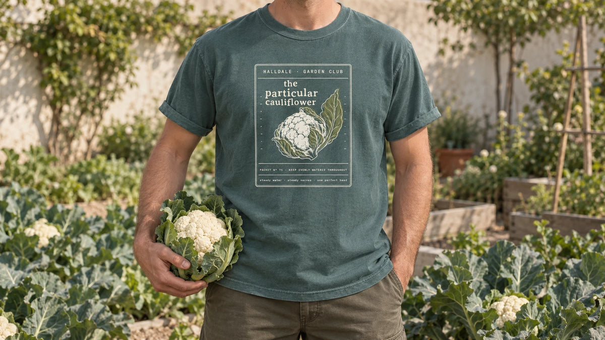 A man in the Blue Spruce Particular Cauliflower tee stands by a brassica bed of cauliflower plants with large leaves, holding one white cauliflower head wrapped in its leaves.