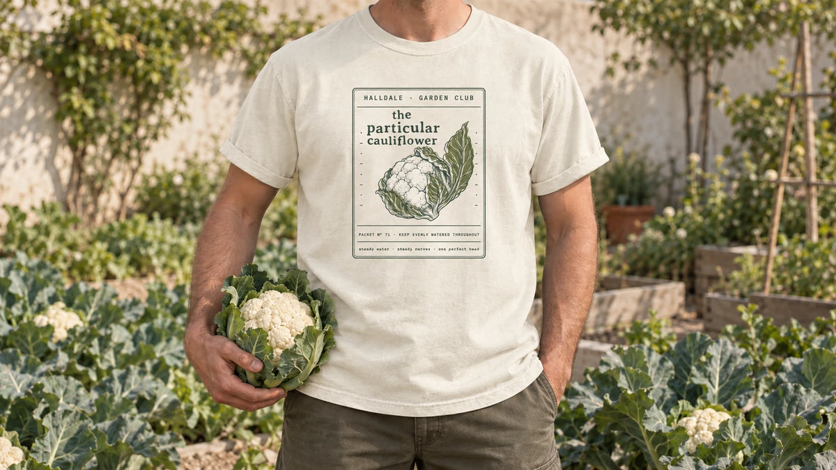 A man in the Ivory Particular Cauliflower tee stands by a brassica bed of cauliflower plants with large leaves, holding one white cauliflower head wrapped in its leaves.