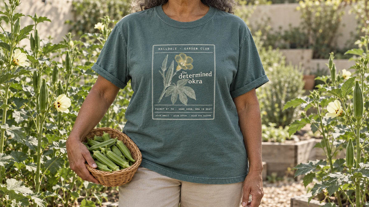 A woman in the Blue Spruce Determined Okra tee stands by tall okra plants with pale yellow flowers and upright pods, holding a small wicker basket of green okra pods.