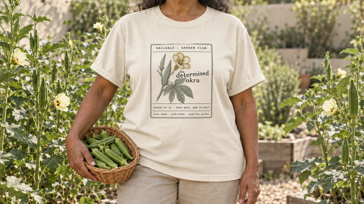 A woman in the Ivory Determined Okra tee stands by tall okra plants with pale yellow flowers and upright pods, holding a small wicker basket of green okra pods.
