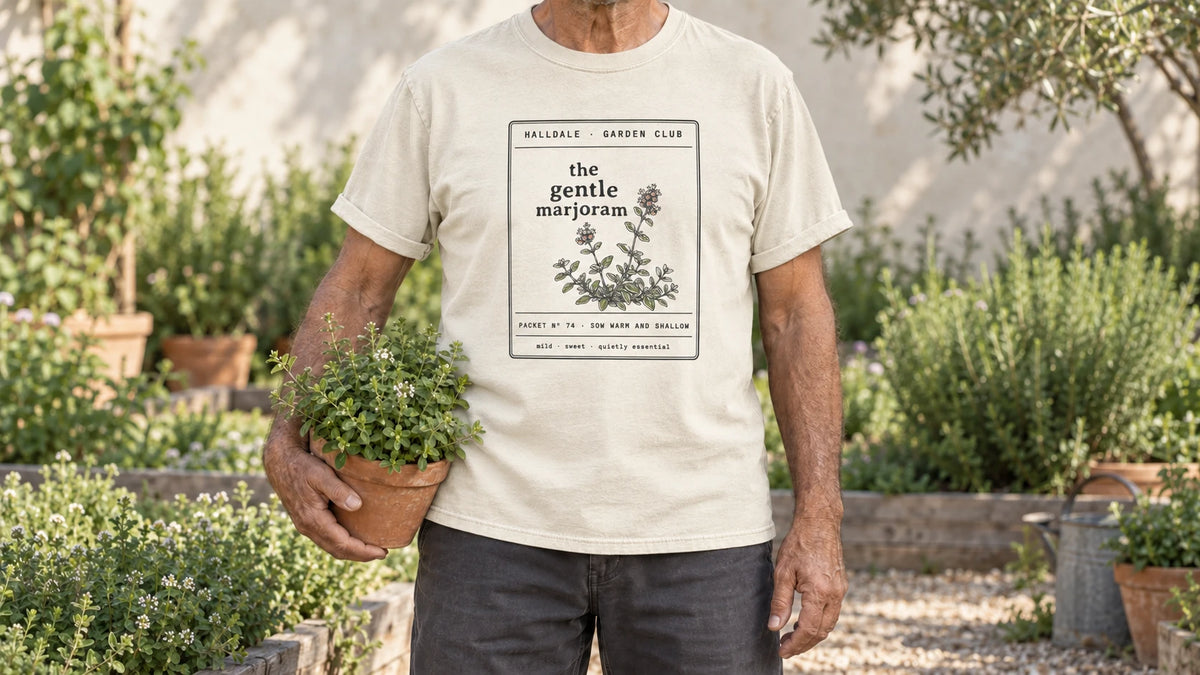 A man in the Ivory Gentle Marjoram tee stands by a sunny herb bed with low mounds of marjoram, holding a terracotta pot of marjoram.