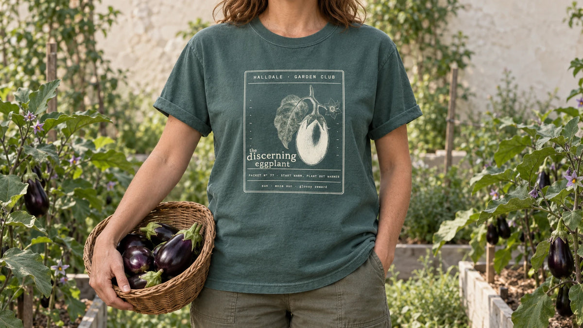 A woman in the Blue Spruce Discerning Eggplant tee stands by eggplant plants with glossy purple fruit and lavender flowers, holding a shallow wicker basket of glossy purple eggplants.