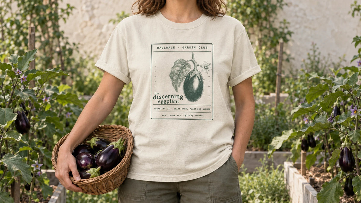 A woman in the Ivory Discerning Eggplant tee stands by eggplant plants with glossy purple fruit and lavender flowers, holding a shallow wicker basket of glossy purple eggplants.