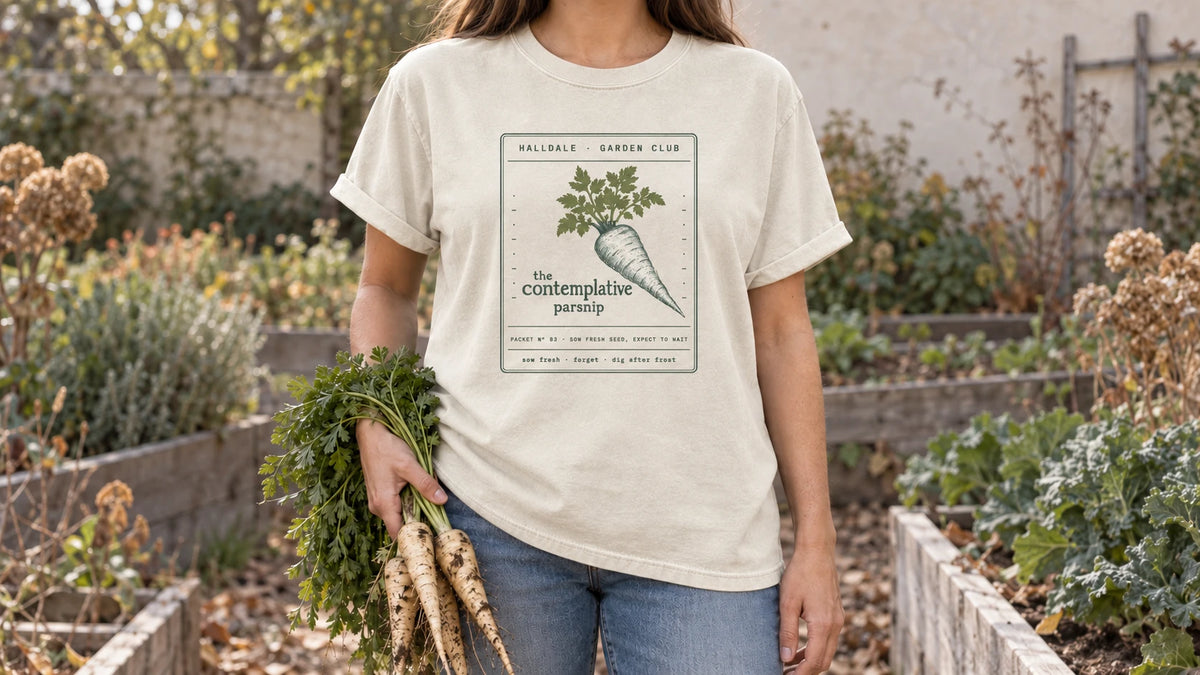 A woman in the Ivory Contemplative Parsnip tee stands by a frosty late fall root bed, holding a bunch of freshly dug parsnips with their leafy tops.